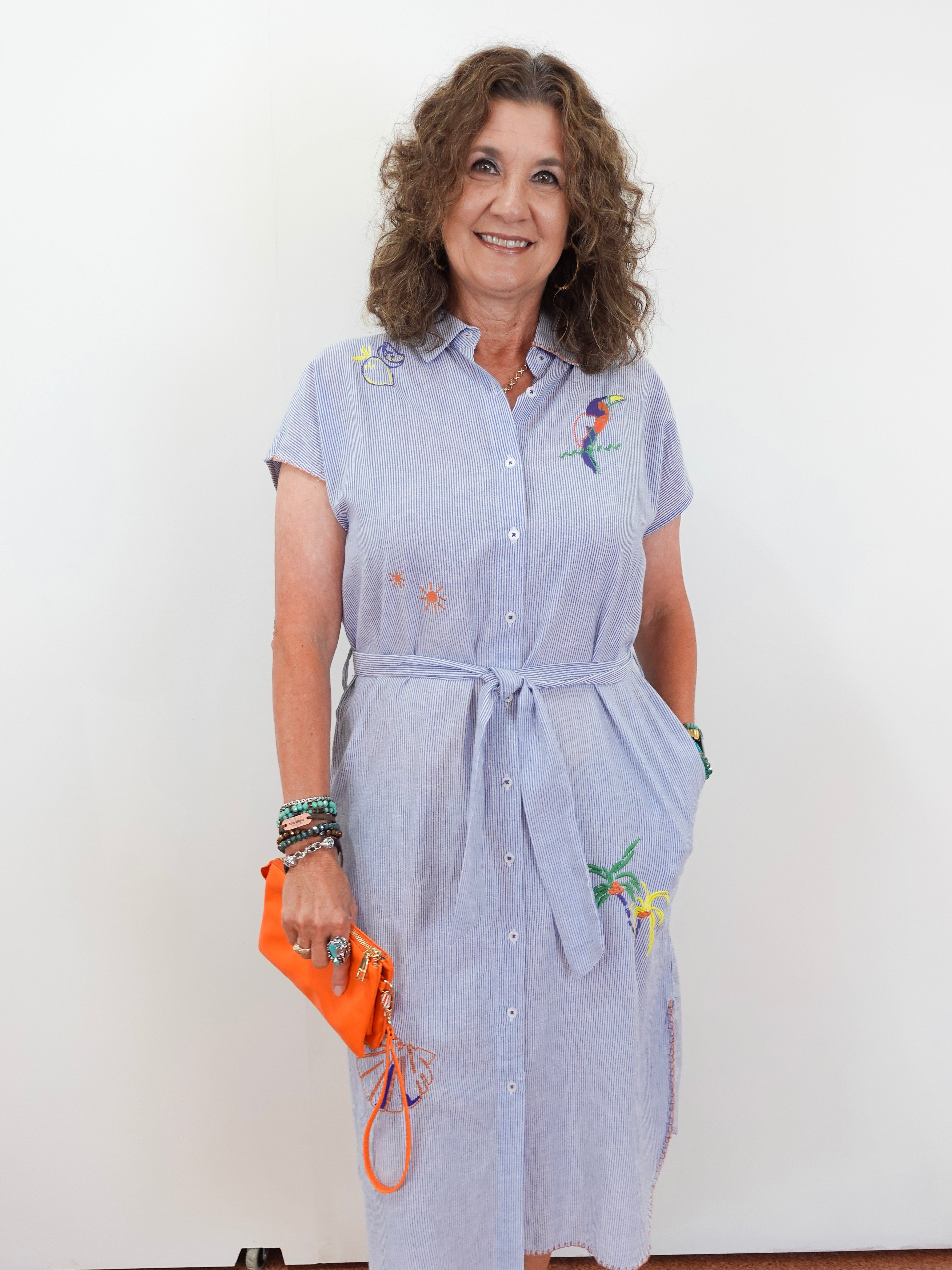 Woman wearing a light blue dress with floral embroidery, holding an orange clutch against a white background