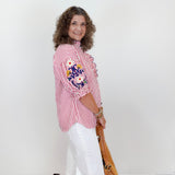 Woman wearing a red and white striped blouse with floral embroidery, white shorts, and holding a brown handbag against a white background.