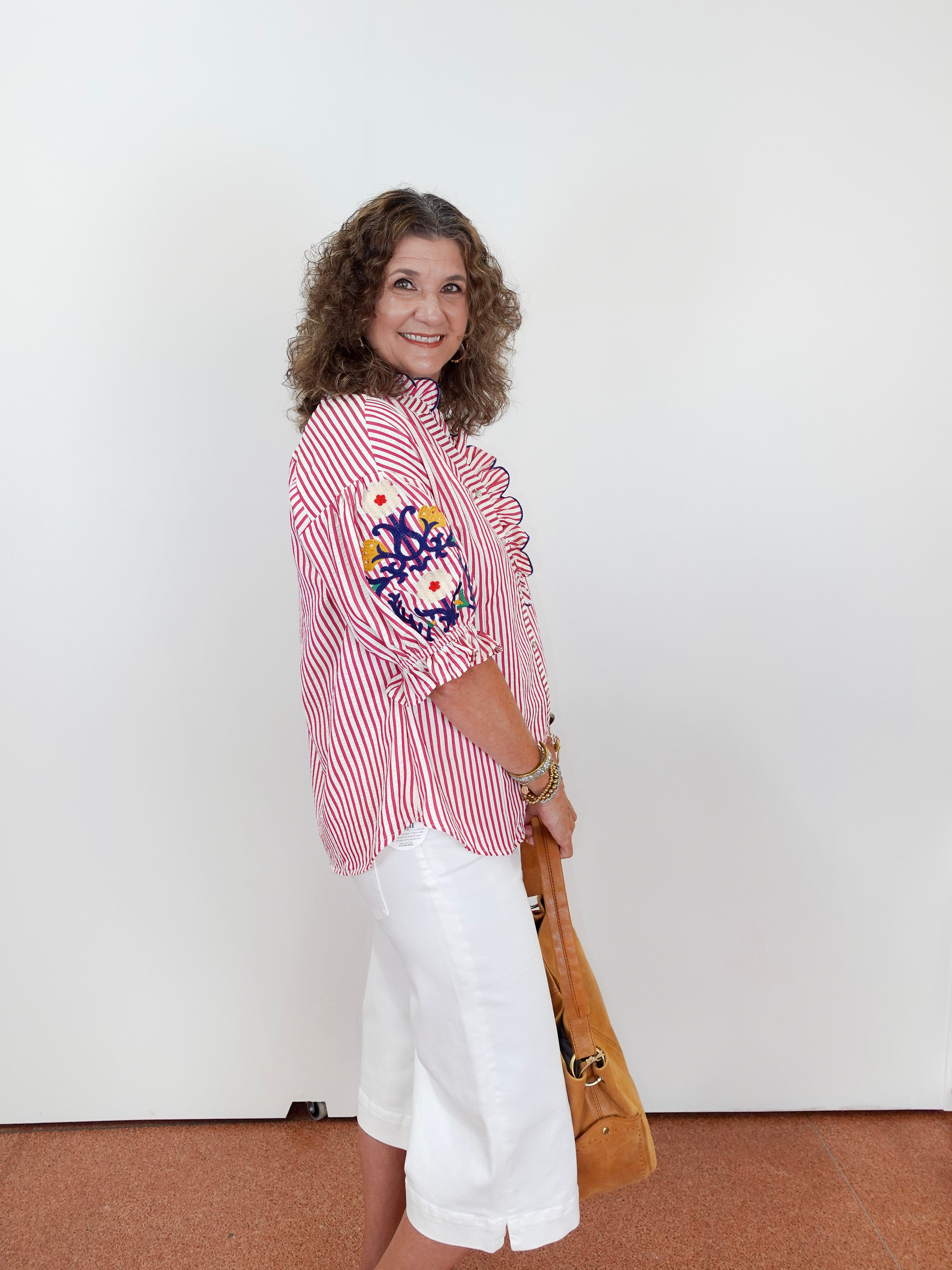 Woman wearing a red and white striped blouse with floral embroidery, white shorts, and holding a brown handbag against a white background.