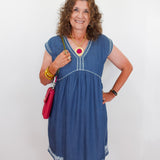 Woman wearing a blue dress with white embroidery, holding a red handbag against a white background.