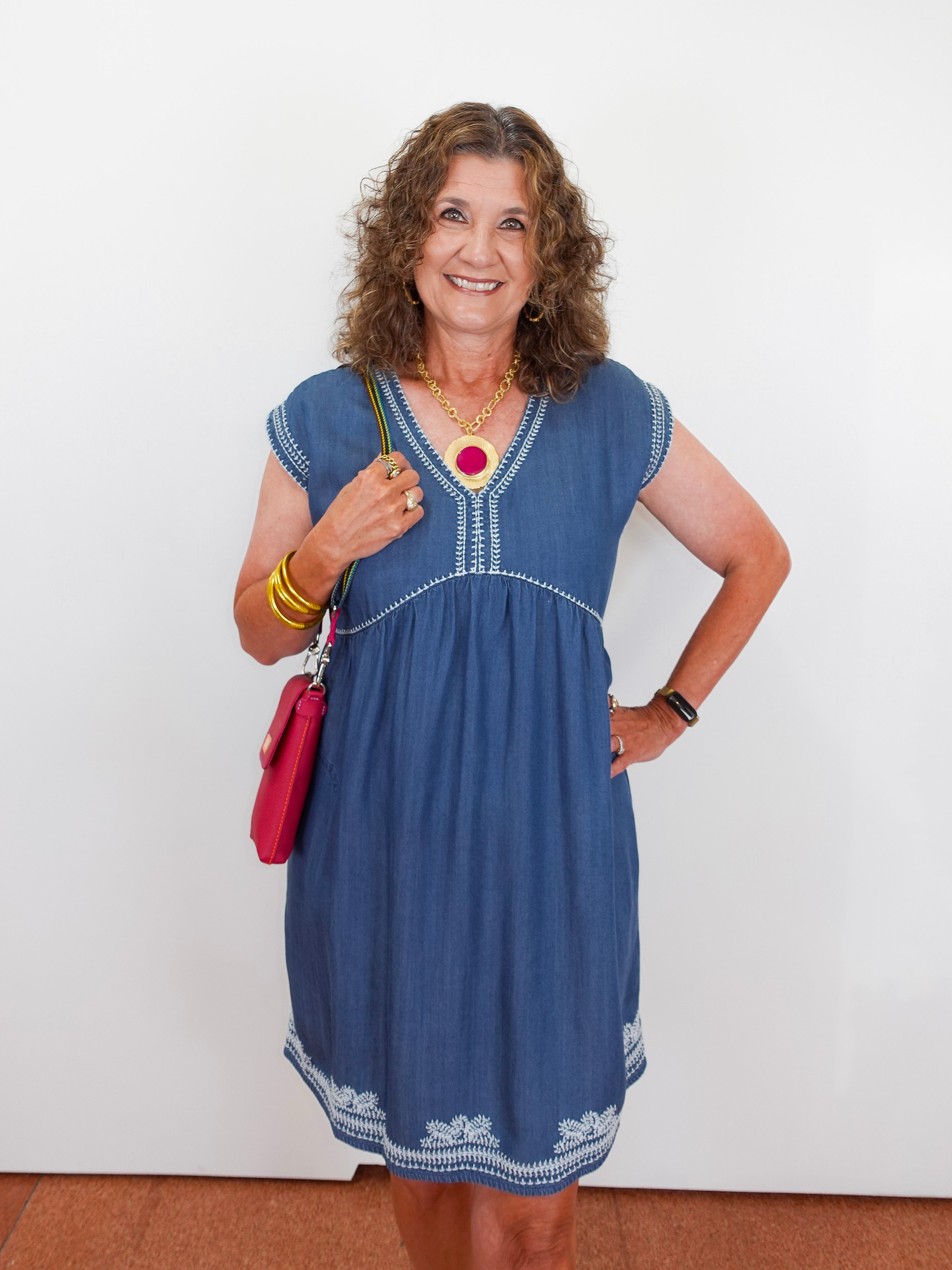Woman wearing a blue dress with white embroidery, holding a red handbag against a white background.