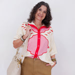 Woman wearing a white blouse with a pink floral design and brown pants, holding a beige handbag against a white background.