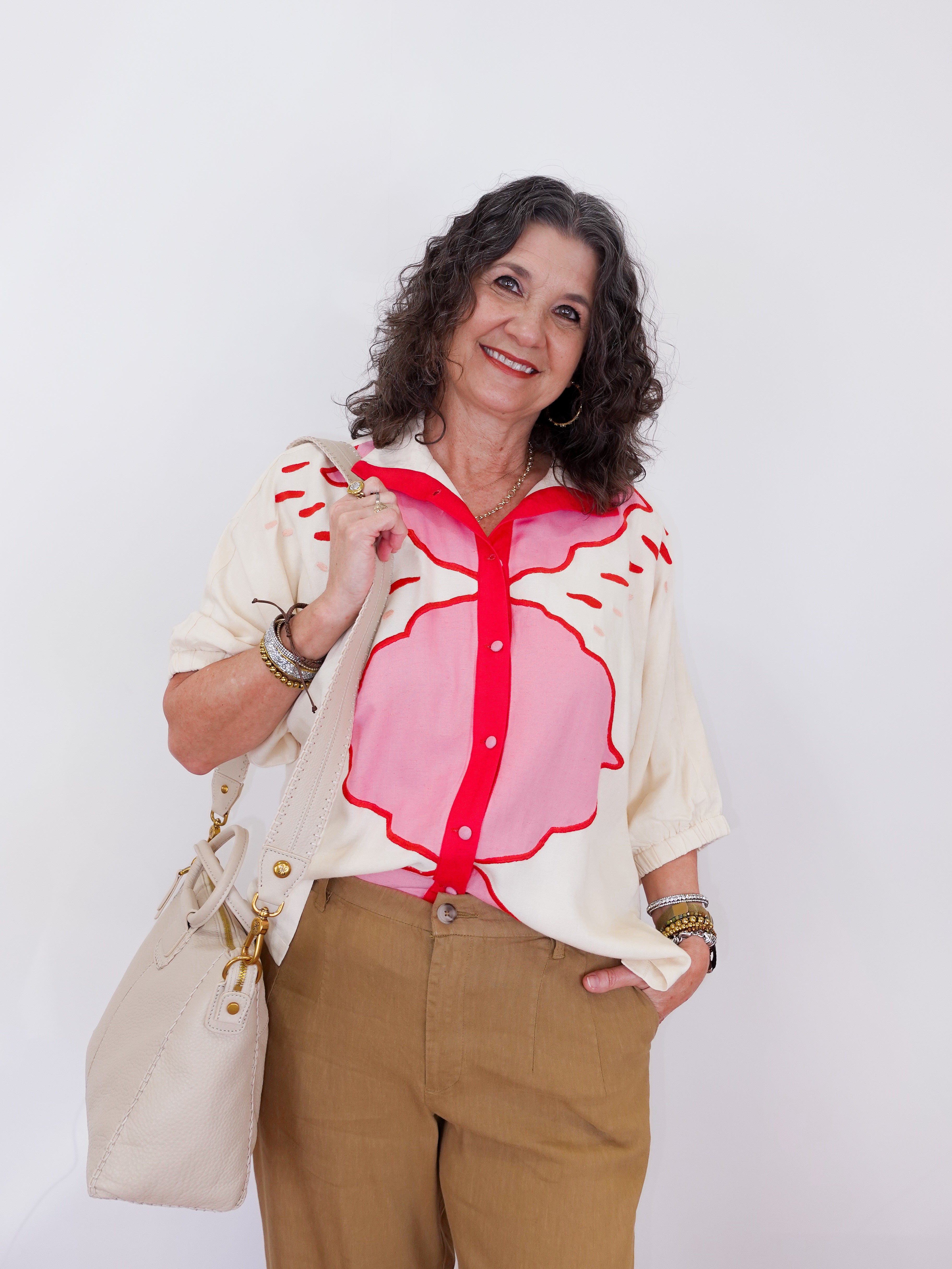 Woman wearing a white blouse with a pink floral design and brown pants, holding a beige handbag against a white background.