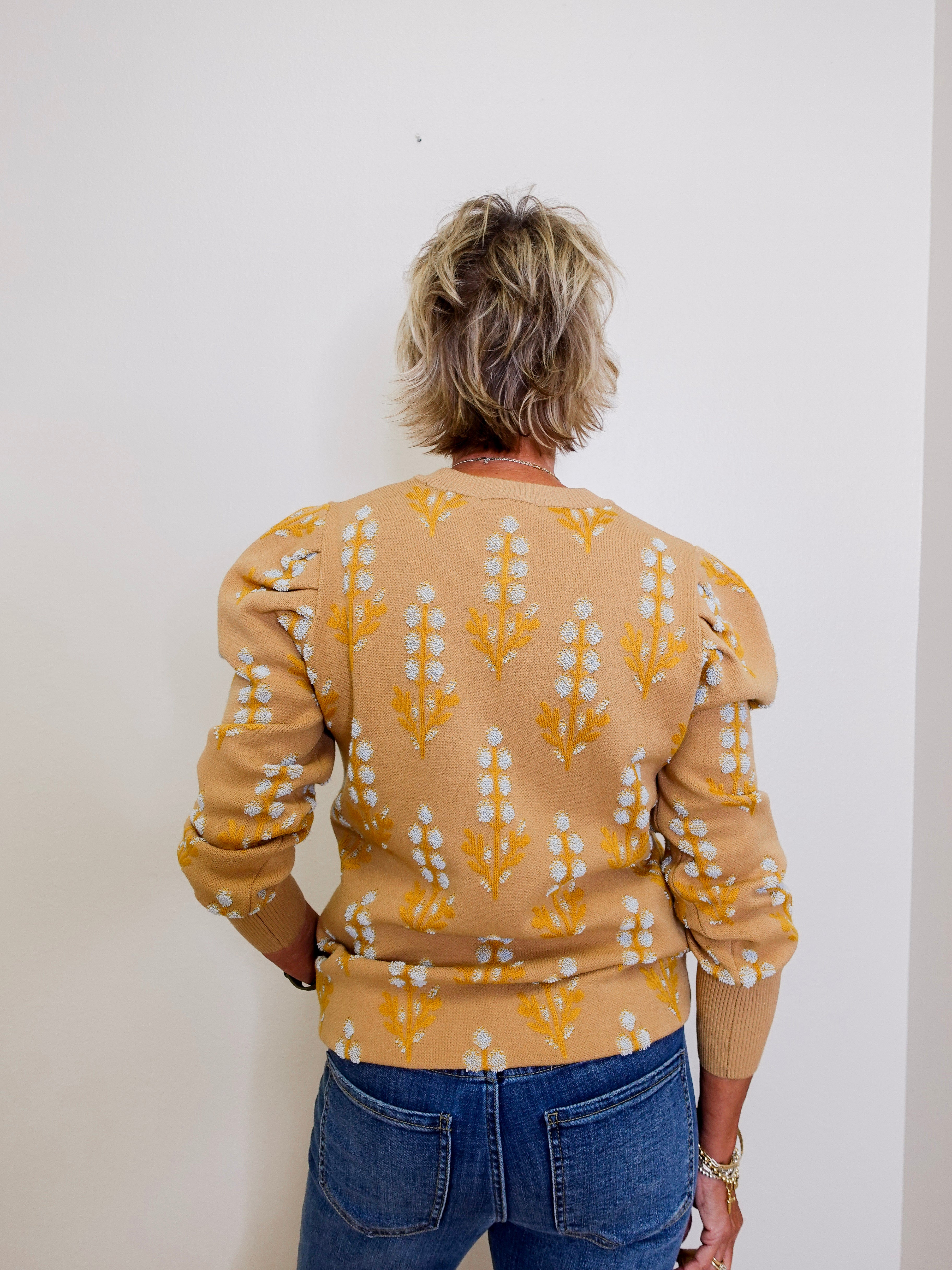 Person wearing a beige sweater with a pattern, standing against a white wall.