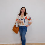 Woman wearing a patterned blouse and jeans, holding a brown handbag against a white wall.