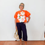 Person wearing an orange floral top with navy pants against a white wall.