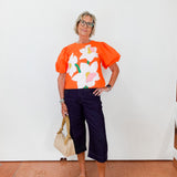Person wearing an orange floral top with navy pants against a white wall.