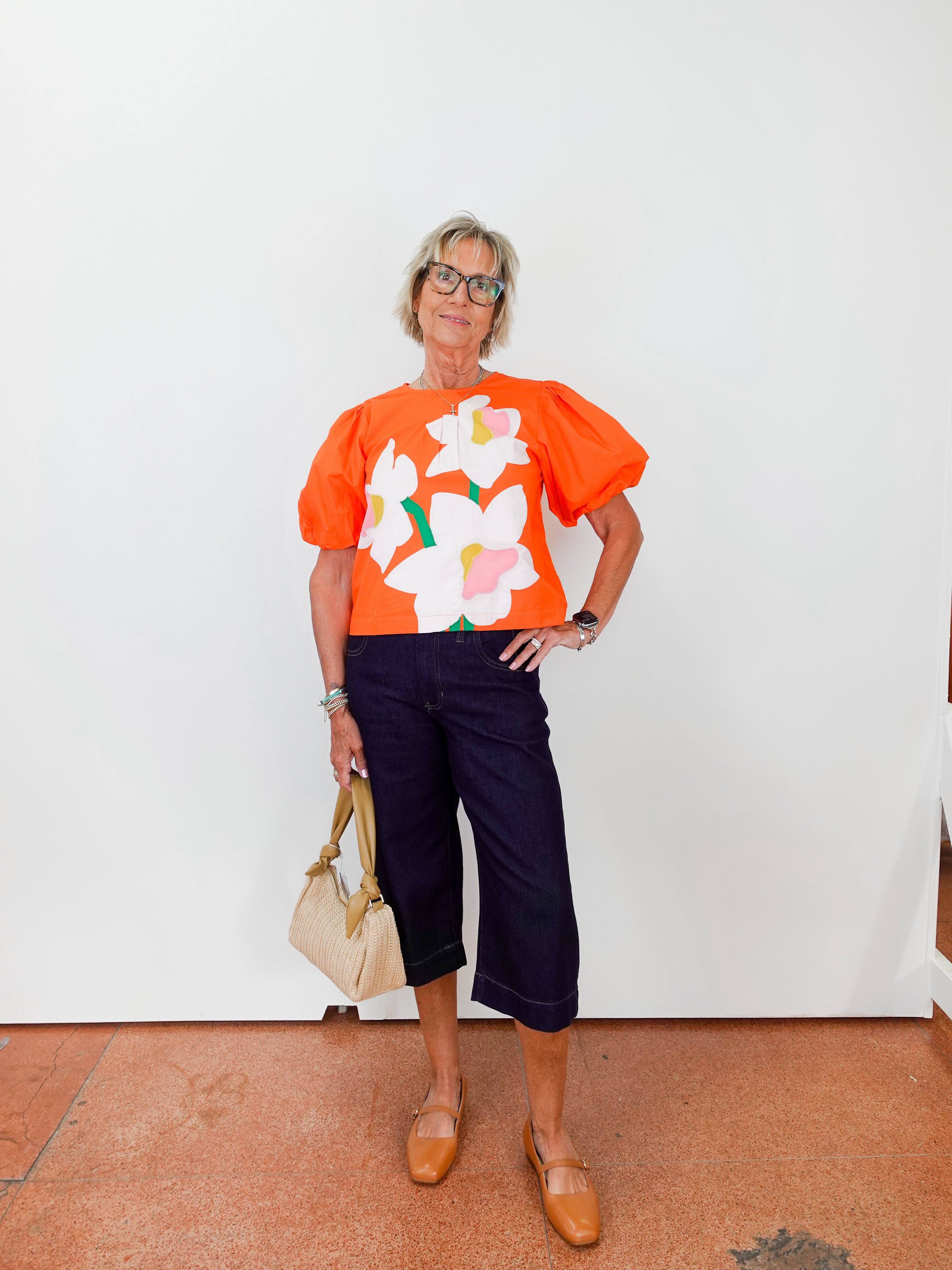 Person wearing an orange floral top with navy pants against a white wall.