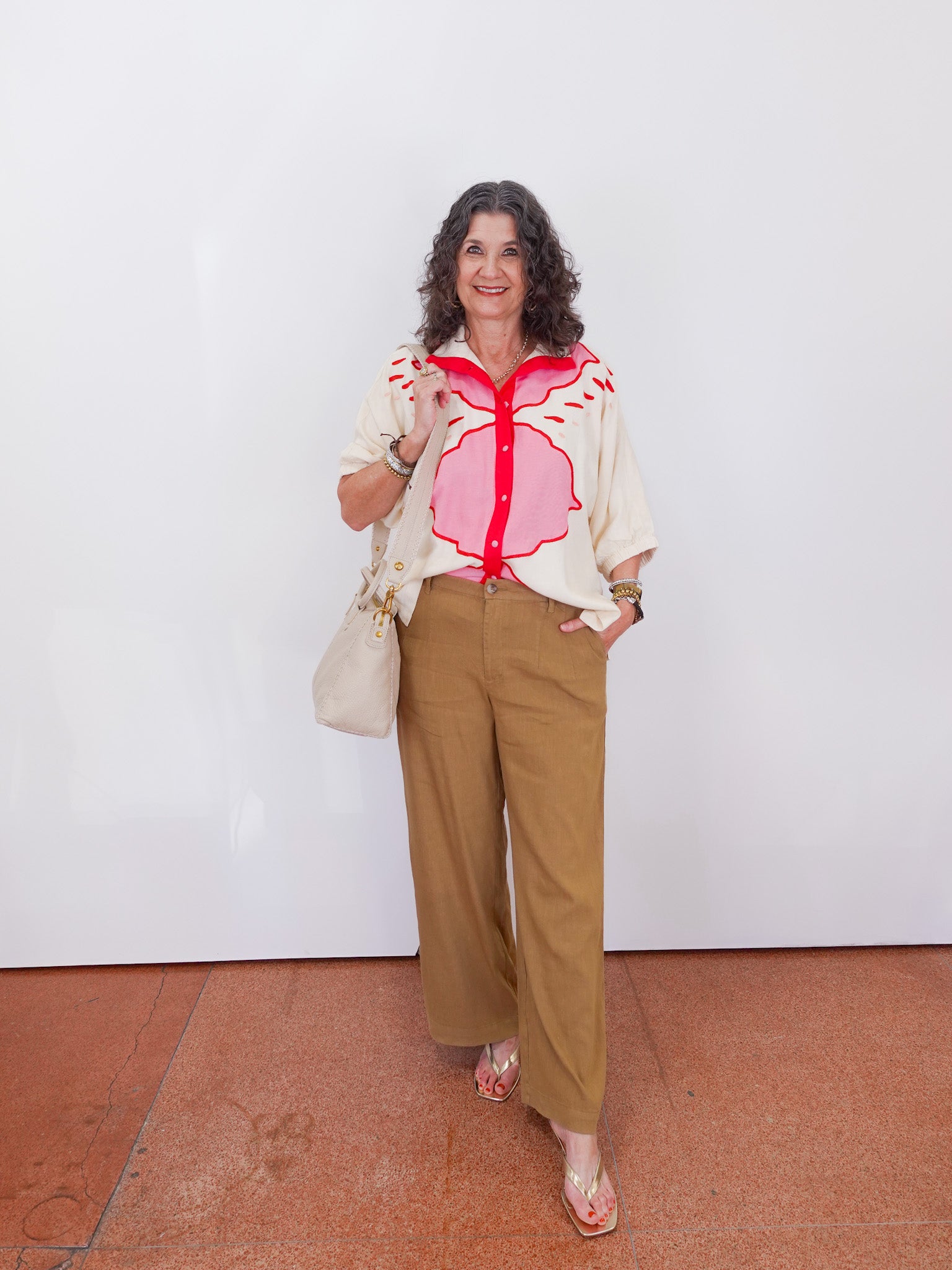 Woman wearing a floral blouse and beige pants standing against a white wall.