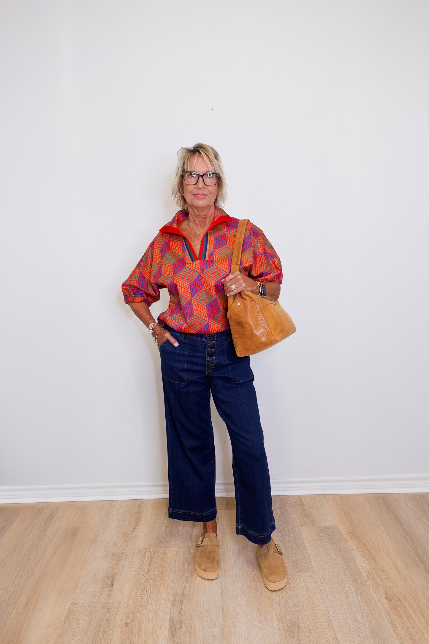 Person wearing a colorful patterned shirt and blue jeans, holding a brown leather bag against a white wall.