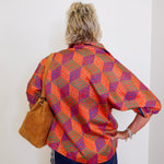 Person wearing a colorful patterned shirt and blue jeans, holding a brown leather bag against a white wall.