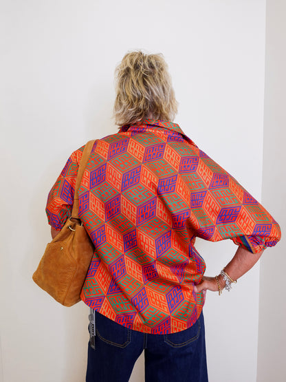 Person wearing a colorful patterned shirt and blue jeans, holding a brown leather bag against a white wall.