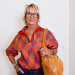 Person wearing a colorful patterned shirt and blue jeans, holding a brown leather bag against a white wall.