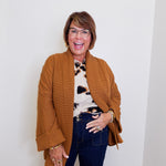 Woman wearing a brown cardigan over a patterned top and dark jeans, standing against a white wall.