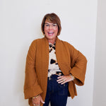 Woman wearing a brown cardigan, patterned top, and dark jeans against a white background
