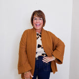 Woman wearing a brown cardigan, patterned top, and dark jeans against a white background