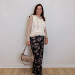 Woman wearing a white lace top and floral pants, holding a beige handbag against a plain background.