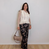 Woman wearing a white lace top and floral pants, holding a beige handbag against a plain background.