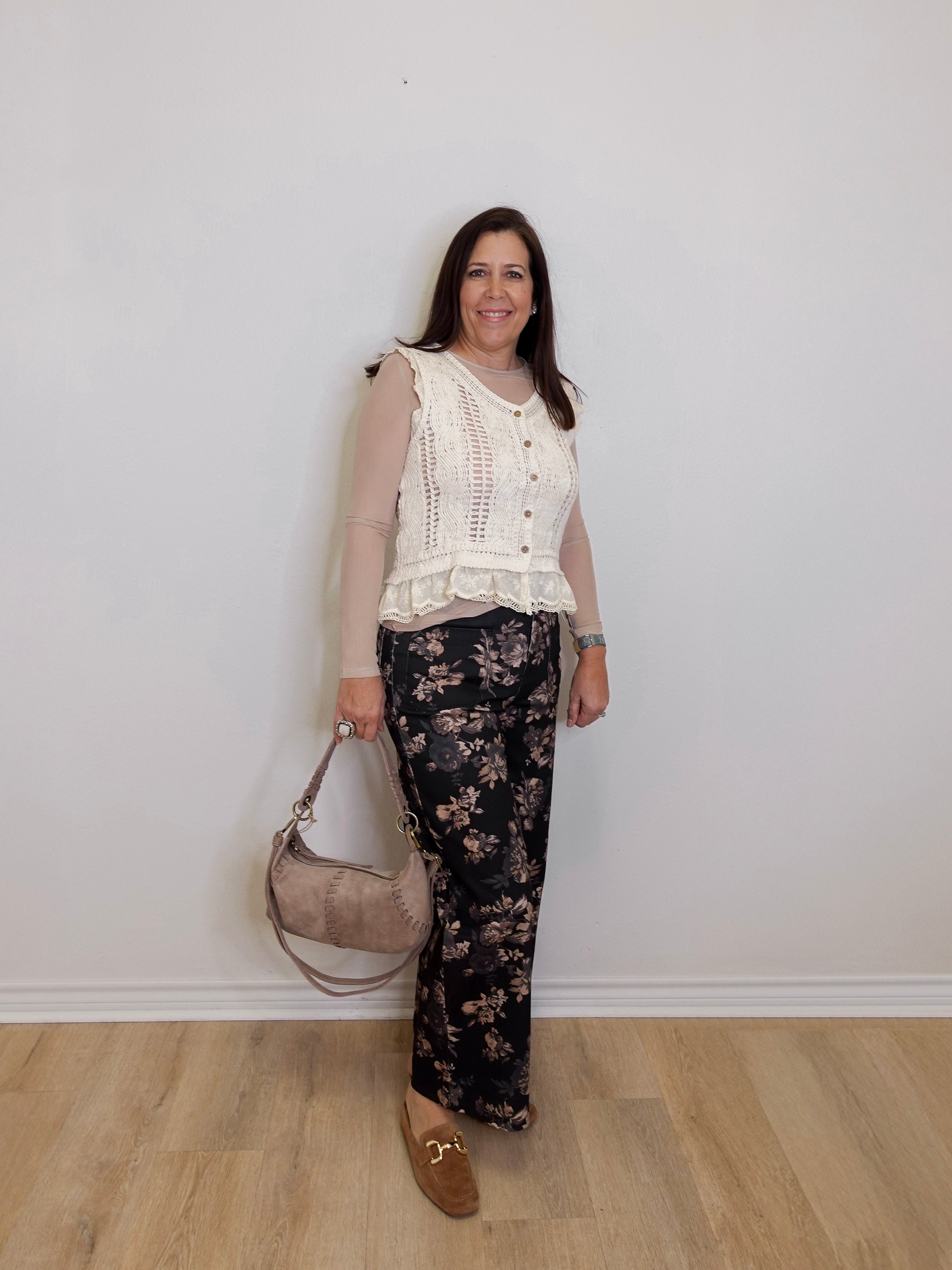 Woman wearing a white lace top and floral pants, holding a beige handbag against a plain background.