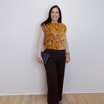 Woman wearing a patterned top and brown pants against a white wall.
