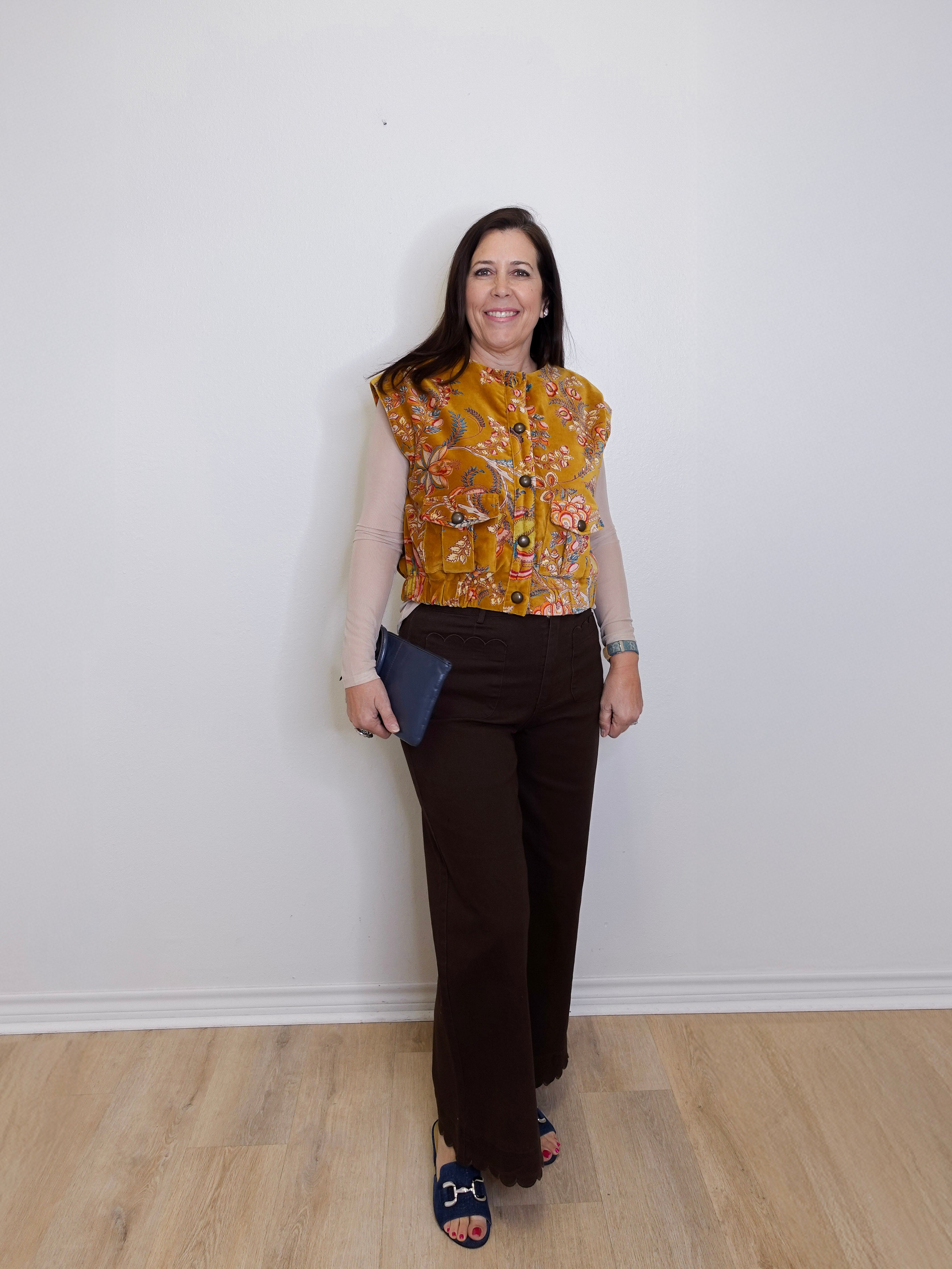 Woman wearing a patterned top and brown pants against a white wall.