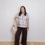 Woman wearing a floral blouse and brown pants against a white wall.