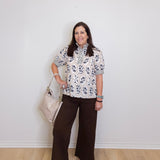 Woman wearing a floral blouse and brown pants against a white wall.