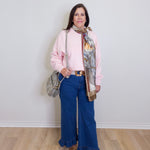Woman wearing a pink sweater, blue jeans, and a patterned scarf against a white wall.