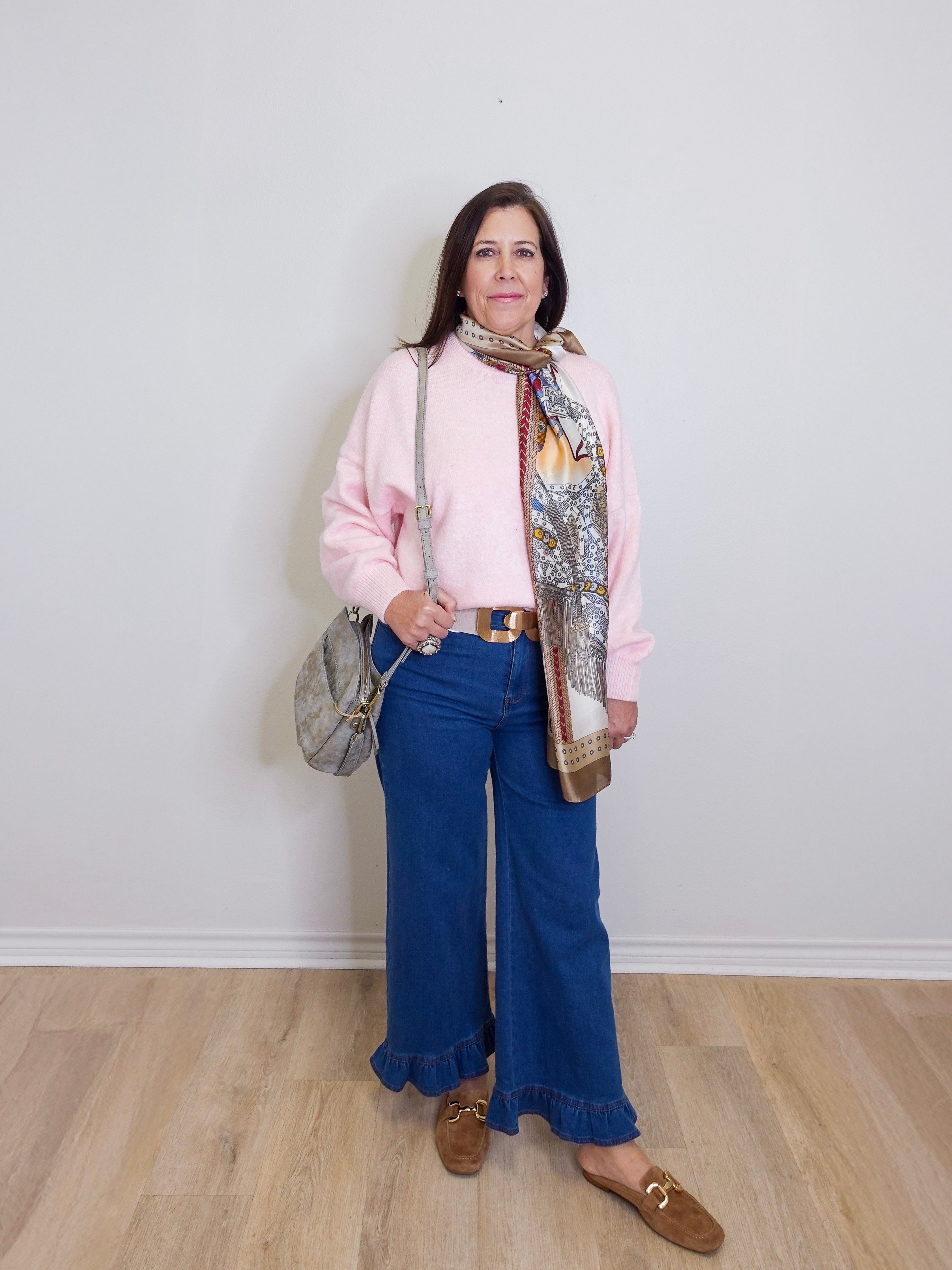 Woman wearing a pink sweater, blue jeans, and a patterned scarf against a white wall.