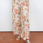 A person wearing wide-leg cropped pants with an allover floral print in peach, pink, and brown on a cream background, paired with beige platform sandals against a plain white wall and terracotta floor.