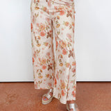 A person wearing wide-leg cropped pants with an allover floral print in peach, pink, and brown on a cream background, paired with beige platform sandals against a plain white wall and terracotta floor.