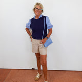 Woman wearing a navy sweater, beige shorts, and brown shoes, holding a blue handbag against a white wall.