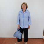 Woman wearing a blue and white striped shirt with black pants, holding a blue handbag against a white wall.