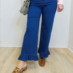 Person wearing blue flared jeans with ruffled bottom hem, pink sweater, and brown loafers on a light wooden floor.