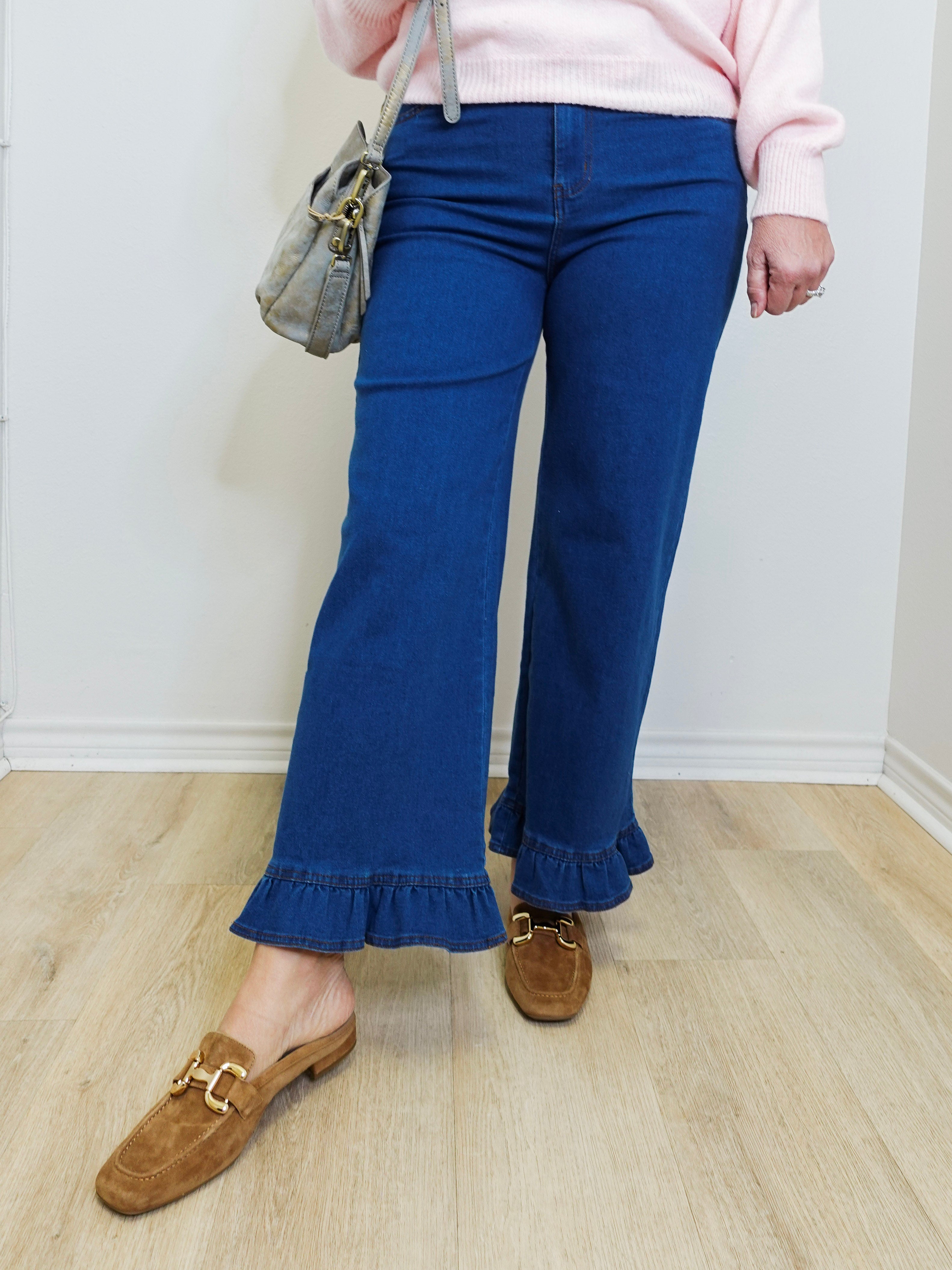 Person wearing blue flared jeans with ruffled bottom hem, pink sweater, and brown loafers on a light wooden floor.