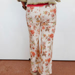 A person shown from the back wearing floral wide-leg pants with a cream base and orange, pink, and green floral print, paired with a beige fringed top and beige wedge sandals against a plain white wall.