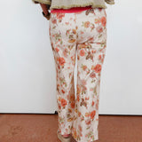 A person shown from the back wearing floral wide-leg pants with a cream base and orange, pink, and green floral print, paired with a beige fringed top and beige wedge sandals against a plain white wall.