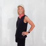 Woman wearing a black sleeveless top and shorts with a pink scarf, standing against a white wall.