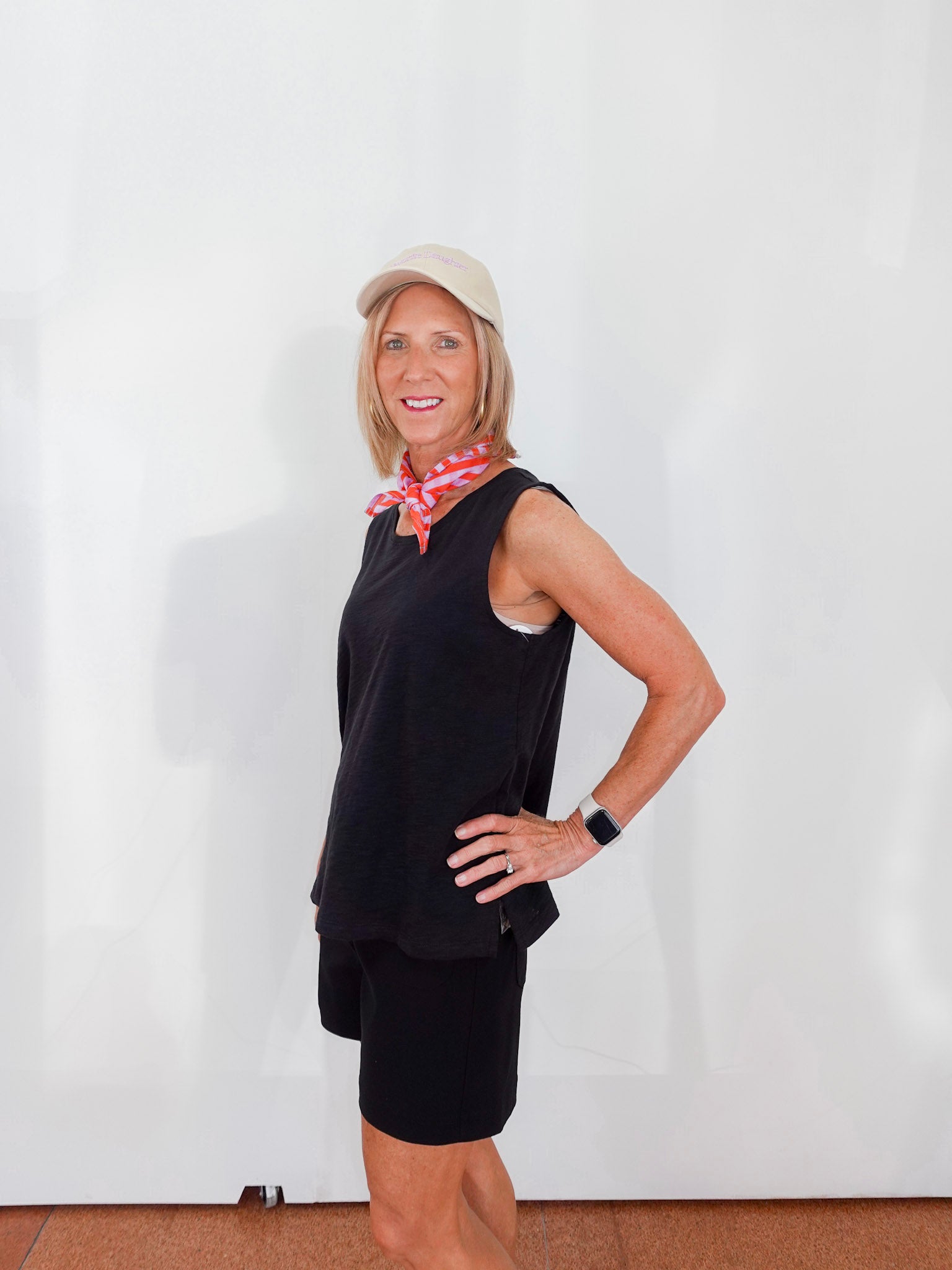 Woman wearing a black sleeveless top and shorts with a pink scarf, standing against a white wall.