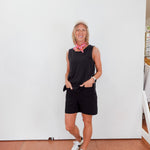 Person wearing a black sleeveless top and shorts with white sneakers, standing against a white wall.