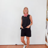 Person wearing a black sleeveless top and shorts with white sneakers, standing against a white wall.