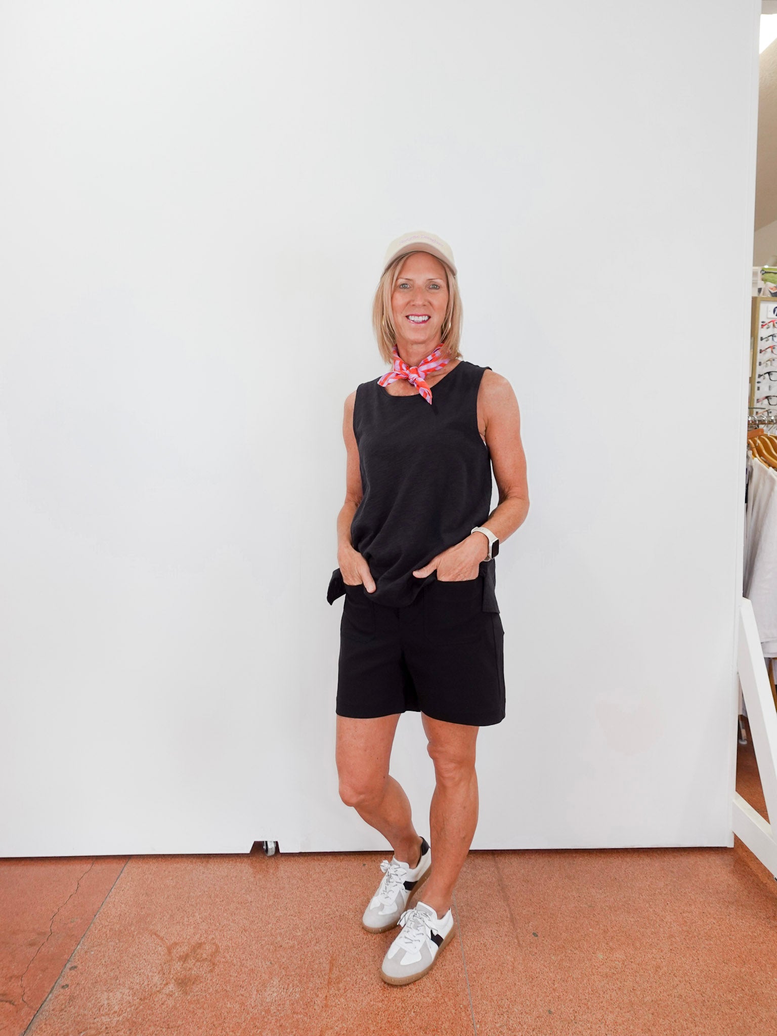 Person wearing a black sleeveless top and shorts with white sneakers, standing against a white wall.
