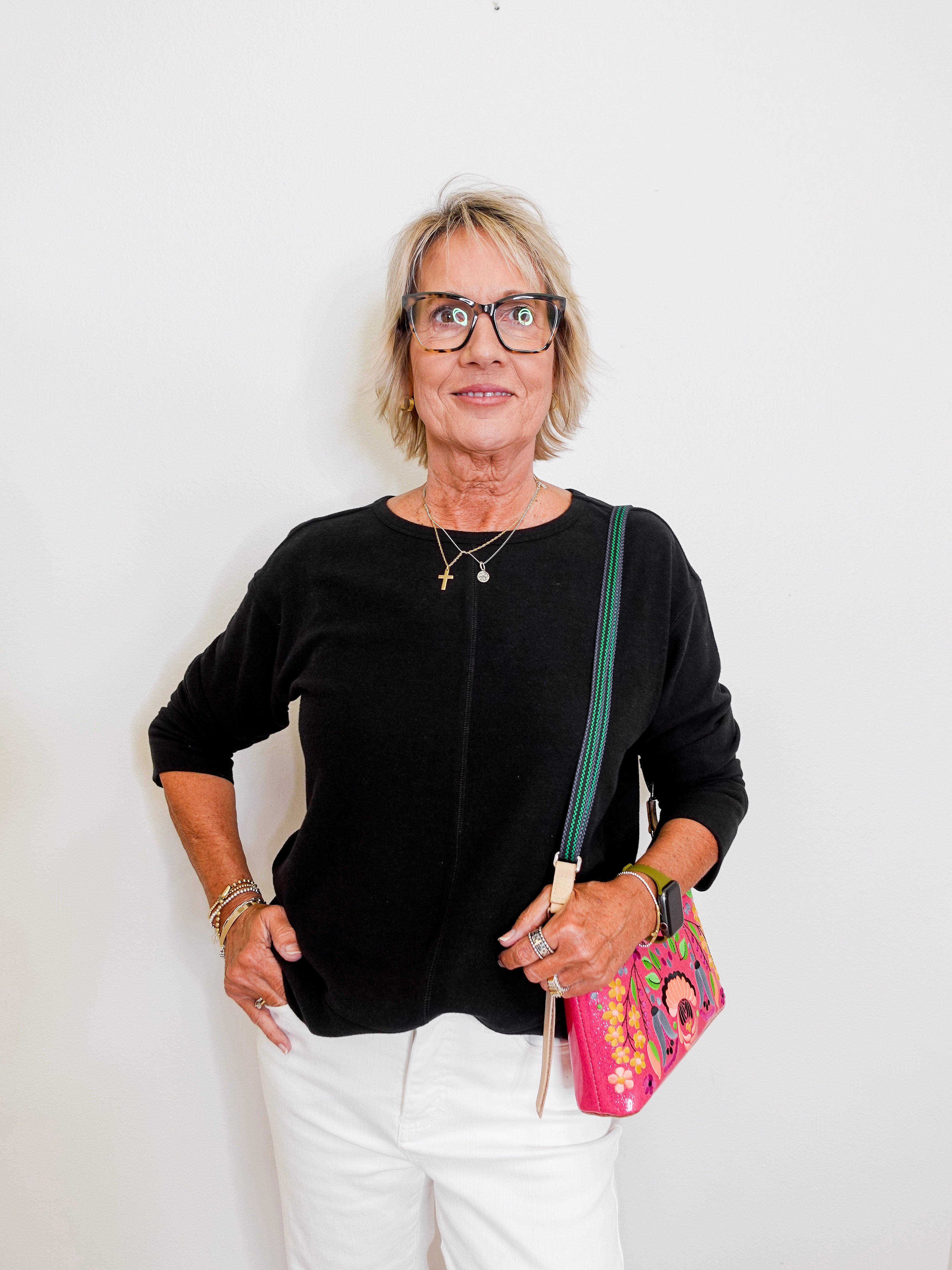 Woman wearing a black top and white pants, holding a colorful bag against a white background
