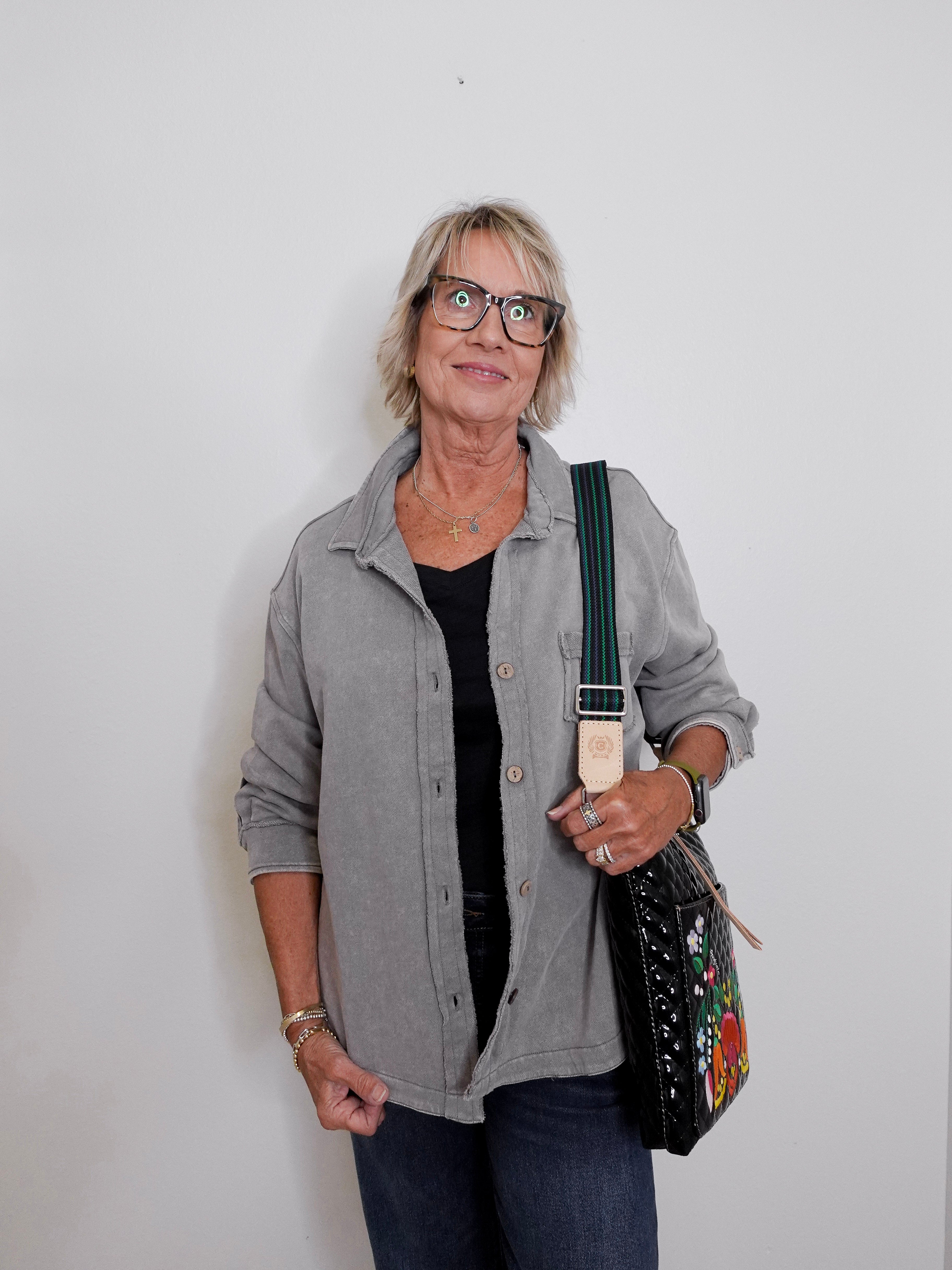 Woman wearing a gray shirt and glasses, holding a black bag with floral patterns against a white background
