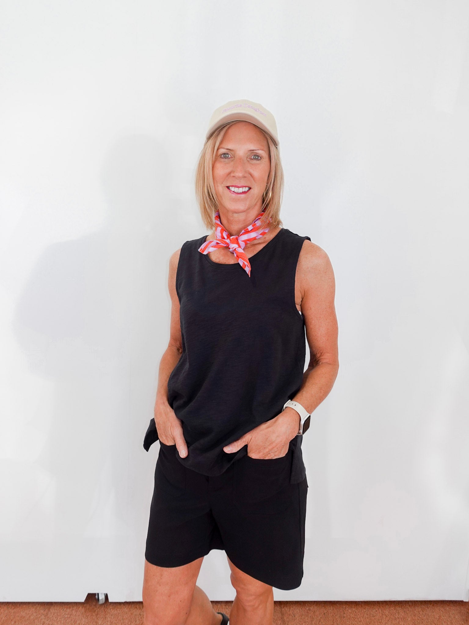 Woman wearing a black sleeveless top and shorts with a red patterned scarf, standing against a white wall.