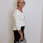 Woman in a white sweater holding a gray handbag against a plain wall.