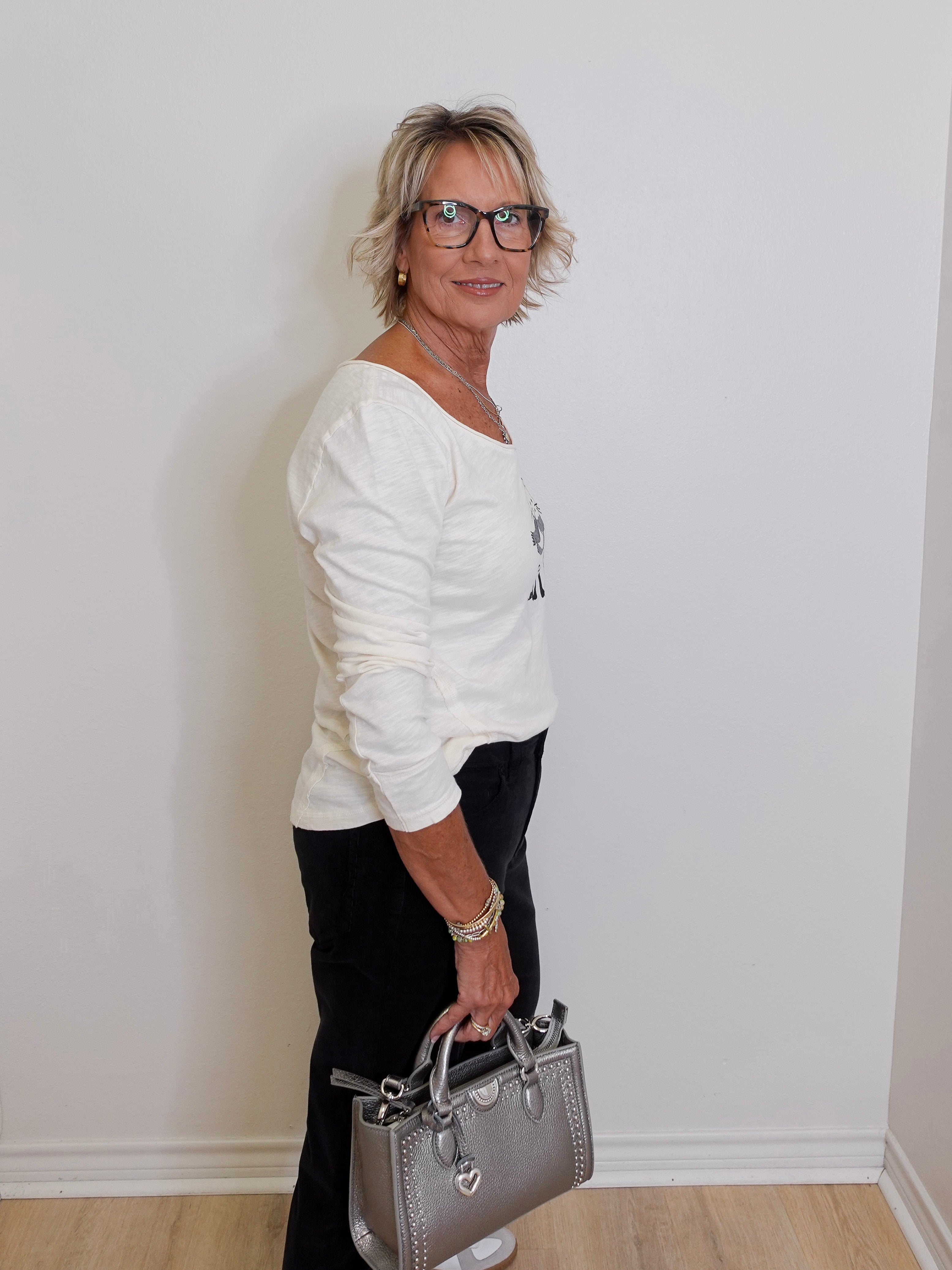 Woman in a white sweater holding a gray handbag against a plain wall.