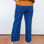 A person shown from the back wearing blue cropped denim jeans, a mustard orange top, and beige heeled sandals against a plain white wall and terracotta floor.