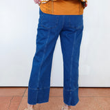 A person shown from the back wearing blue cropped denim jeans, a mustard orange top, and beige heeled sandals against a plain white wall and terracotta floor.