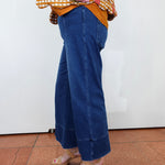 A person wearing blue wide-leg cropped jeans with rolled cuffs, paired with a yellow-and-white gingham top and beige heeled sandals, standing against a plain white wall on a terracotta tiled floor.